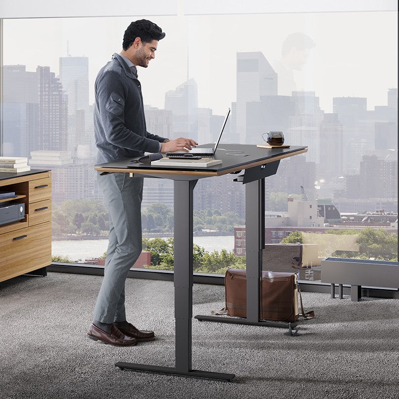 BDi Sequel 20 Sit + Stand Desk in Natural Walnut, raised, with a person standing at it, beside a multi-function cabinet
