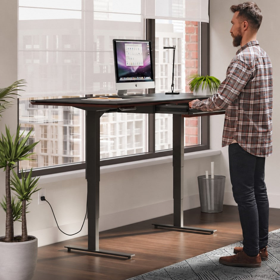 BDi Sequel 20 Sit + Stand Desk in Chocolate Stained Walnut, raised, with a person standing at it, beside a multi-function cabinet