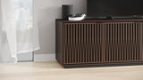 BDi Elements 8703 Media + Storage Cabinet in Toasted Oak, with a TV and a speaker, by a window, with Tune door fronts