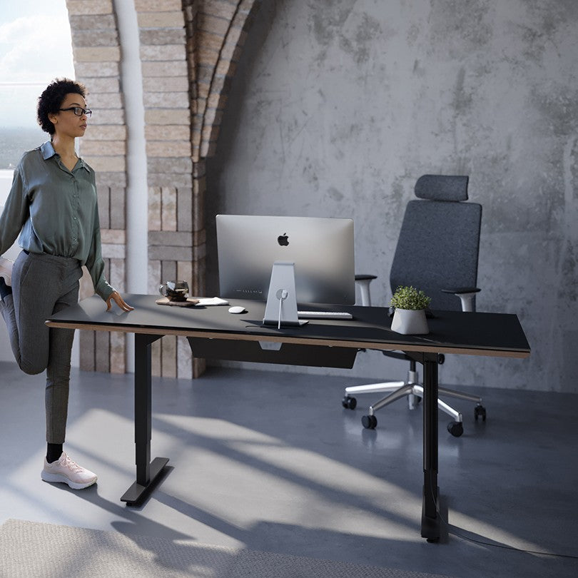 BDi Sequel 20 Sit + Stand Desk in Natural Walnut, with a person stretching beside it, in a loft